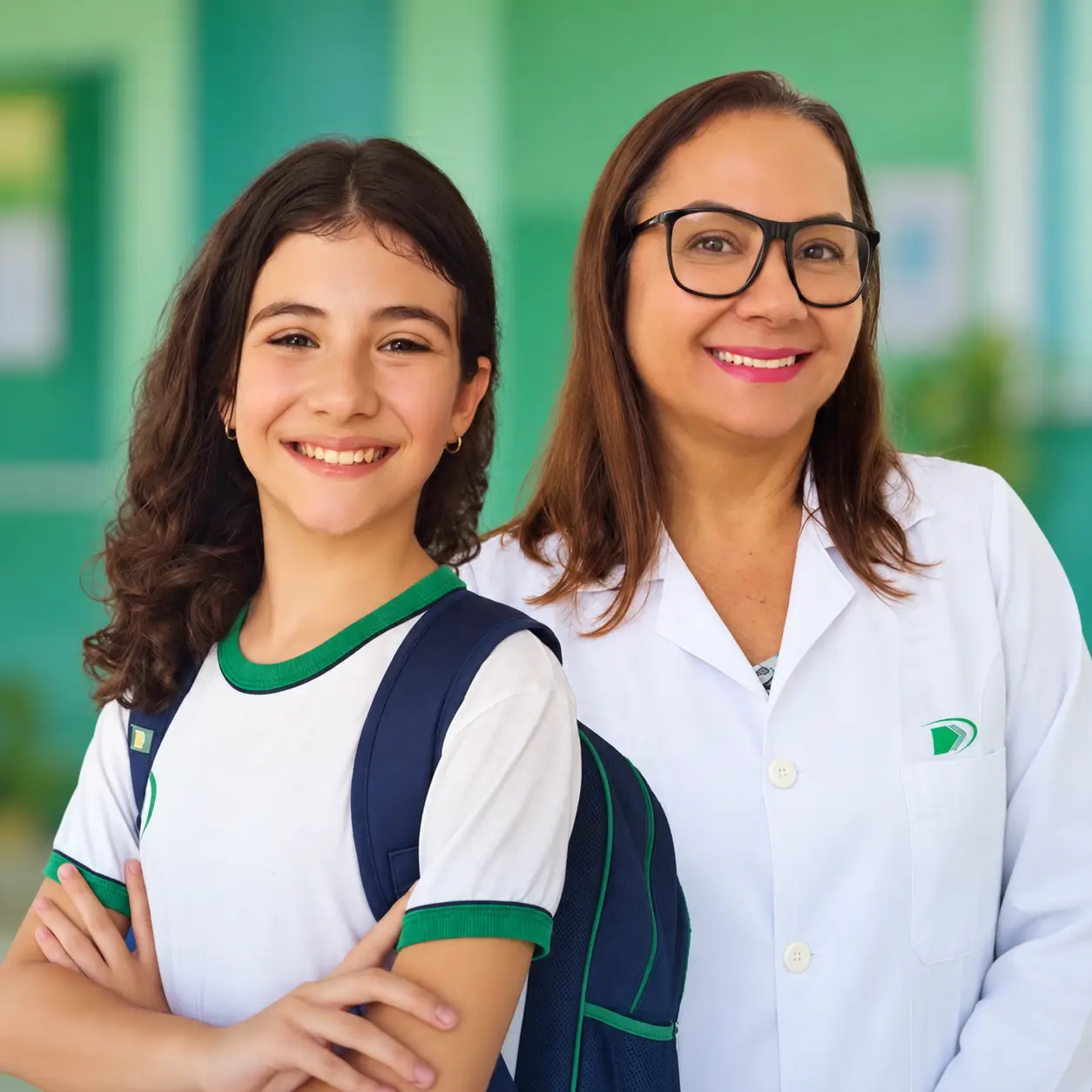 Aluna do Colégio Domum, de mochila e uniforme, ao lado de uma professora de jaleco, as duas sorrindo